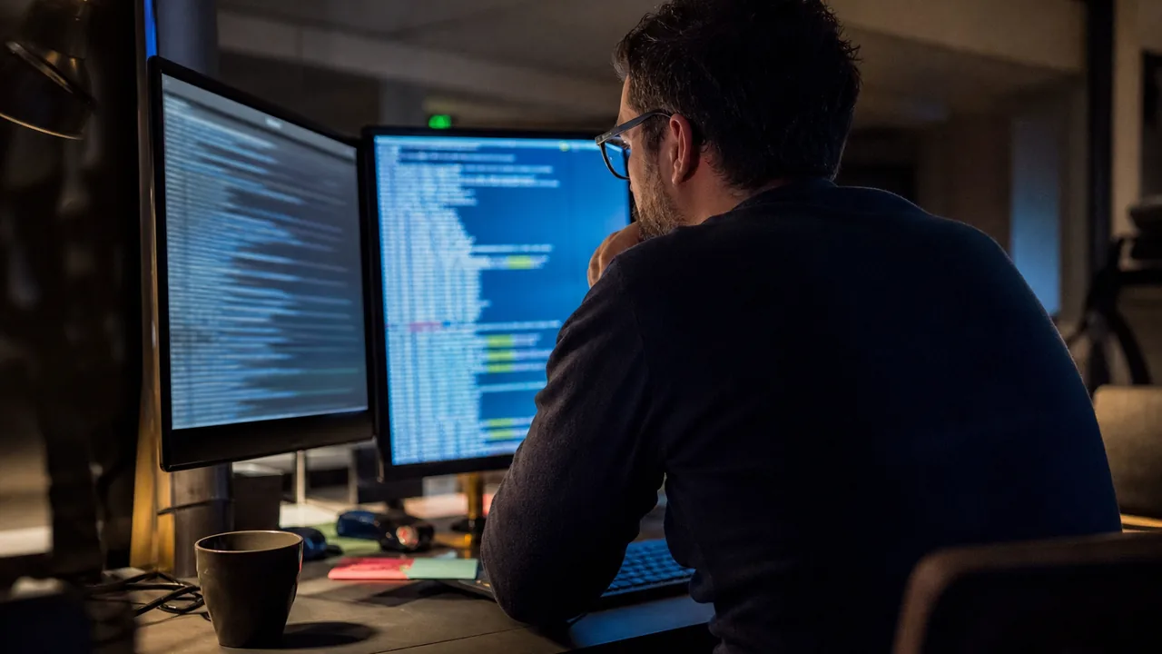 Engineer on a late shift reading deployment logs on two monitors in a small office